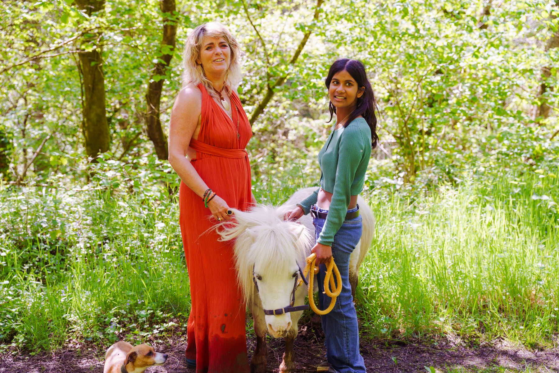 Carolyn in a rust-coloured dress standing with a small white pony in dappled woodland